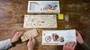A wooden table with a Ugears 3D mechanical wooden puzzle VM-02 motorcycle kit, including the box, unassembled parts, and instructions. Two people are holding parts of the kit. Anteprima dell’immagine prodotto