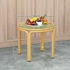 A round rattan side table with a glass top, holding a fruit platter. The table is placed on a textured floor in front of a wood-paneled wall. Podgląd zdjęcia produktu