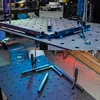 Two images show metal square tubes being clamped onto a perforated welding table, with some tubes already welded into an L-shape. The top image shows a larger scale setup with more equipment in the background, while the bottom image is a closer view of the Náhled obrázku produktu
