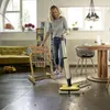 A woman is using a floor cleaner in a living room with furniture and shelves. Produktbild-Vorschau