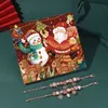 A Christmas advent calendar with a festive design, featuring Santa Claus and a snowman, is placed on a dark green surface. Two charm bracelets are laid out in front of the calendar. Produktbild-Vorschau