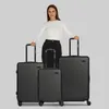 A woman stands behind three black suitcases of varying sizes against a plain gray background. Produktbild-Vorschau