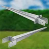Two galvanized steel post anchors are shown against a blurred background of green foliage and blue sky. Náhled obrázku produktu