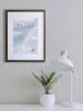 A framed picture hangs on a wall above a desk with a lamp and a potted plant. Produktbild-Vorschau