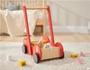 A red wooden walker with blocks inside, placed on a carpeted floor next to a bed. Produktbild-Vorschau