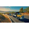 Several dump trucks driving on a winding road with a city skyline in the background. Náhľad obrázku produktu