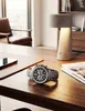 A watch is placed on a wooden desk next to a lamp and a book. Anteprima dell’immagine prodotto