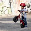 A young boy wearing a red helmet rides a red balance bike in front of a graffiti-covered wall. Produktbild-Vorschau