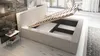 A bed with its storage compartment open, showing the slatted base and internal storage space. A small side table with decorative items is next to the bed. Produktbild-Vorschau