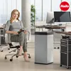 A woman sits in an office chair holding a magazine next to a silver trash can. Produktbild-Vorschau