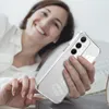 A woman is lying in bed, smiling and holding a clear phone case with a Samsung phone inside. Produktbild-Vorschau