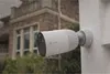 A security camera mounted on a wall during a rain shower, with a house in the background. Produktbild-Vorschau