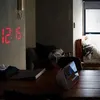 A bedside table with a digital clock and a bed in the background. Produktbild-Vorschau