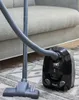 A black vacuum cleaner with a gray hose and nozzle is on a patterned rug in front of a gray sofa. Produktbild-Vorschau