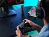 A person wearing headphones is playing a video game using a controller, with a computer monitor and a drink can on the desk. Náhled obrázku produktu