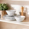 A set of white ceramic bowls and plates with a black abstract design, arranged on a wooden shelf next to a plant and a pepper grinder. Náhled obrázku produktu