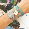 A close-up of a person's wrist wearing a silver Casio watch and several blue string bracelets, one with a cowrie shell charm. In the background, out of focus, are pink flowers and greenery. Podgląd zdjęcia produktu