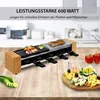 A raclette grill with food and cheese platter on a wooden table, with kitchen appliances in the background. Produktbild-Vorschau