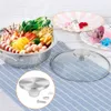 A stainless steel pot lid knob and screws are displayed on a blue and white checkered cloth, next to a glass pot lid. In the background, a pot filled with food and decorative plates are visible. Produktbild-Vorschau