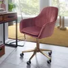 A pink velvet office chair with a gold base and wheels is placed next to a wooden desk with drawers. The scene is set in a room with a yellow rug and a large window in the background. Produktbild-Vorschau