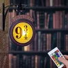 A Harry Potter themed night light shaped like the Platform 9 3/4 sign, mounted on a wall with a bookshelf in the background. A hand is holding a remote control to change the light's color. Produktbild-Vorschau