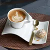 A white porcelain cup of coffee with latte art sits on a matching saucer, accompanied by a gold spoon and a small pastry, all placed on a wooden tray. Produktbild-Vorschau