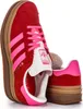 A close-up, angled view of two Adidas Gazelle Bold women's sneakers in red and pink, with a stone placed between them. Produktbild-Vorschau