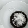 A close-up view of salt crystals in a small dark bowl, placed on a light-colored plate. Produktbild-Vorschau