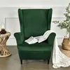 A green wingback armchair with a blanket and newspaper on it, next to a side table with a lamp and a plant in a basket. Anteprima dell’immagine prodotto