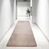 A long, rectangular rug is laid out in the middle of a modern, white hallway. The hallway has white walls and white tiled floors. There is a plant in a pot in the background. Náhled obrázku produktu