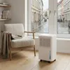 A white dehumidifier is placed on a wooden floor next to a beige armchair, with a blanket draped over it. The scene is set indoors, with a large window showing a rainy street view. Podgląd zdjęcia produktu