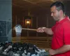 A man in a red shirt is pouring water from a ladle onto hot stones in a sauna. Produktbild-Vorschau