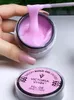 A hand holding a spatula with pink gel for nails, with a jar of the gel in the background. Náhled obrázku produktu