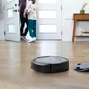 A robot vacuum cleaner is on a wooden floor in a hallway, with two people walking through a doorway in the background. Voir les images du produit