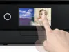 A finger is touching the screen of a black printer, displaying two images side-by-side: a landscape and a baby. Náhled obrázku produktu