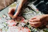 A person's hands are shown painting a canvas with a brush and small paint pots. The canvas has a floral pattern with numbers indicating where to paint. Produktbild-Vorschau