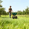 A man is pushing a fertilizer spreader through a grassy field with trees in the background. Produktbild-Vorschau
