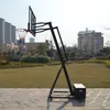A portable basketball hoop stands on a paved area in front of a grassy field and buildings. Produktbild-Vorschau