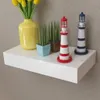 A white floating shelf with decorative items including a yellow vase with yellow flowers, two small potted plants, and two lighthouse figurines. Náhľad obrázku produktu