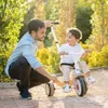 A father is helping his young son ride a beige tricycle in an outdoor setting with trees and sunlight. Produktbild-Vorschau