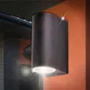 A modern outdoor wall lamp is mounted on an orange wall, with a dark roof and brick wall visible in the background. Produktbild-Vorschau