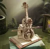 A wooden mechanical cello model is displayed on a stack of books, with a blurred background of greenery and a framed picture. Voir les images du produit