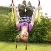 A young girl is hanging upside down from a wooden swing set with yellow rings in a grassy outdoor area. Produktbild-Vorschau