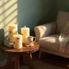 Three LED candles with a birch wood look are placed on a wooden side table next to a mug of coffee. A chair with a knitted blanket is visible in the background. Produktbild-Vorschau