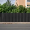 A black corrugated metal fence in front of green trees and a building. Náhled obrázku produktu