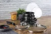 A set of black pots and pans with glass lids, a detachable handle, a jar of pasta, a small bowl of chili peppers, and a potted plant are arranged on a wooden surface against a brick-patterned wall. Podgląd zdjęcia produktu