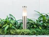 A modern outdoor lamp is placed in a garden bed with lush green plants in front of a white wall. Anteprima dell’immagine prodotto