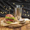 A burger and a stainless steel cooking pot are displayed on a wooden board with fairy lights in the background. Produktbild-Vorschau