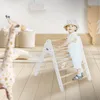 A baby is climbing on a wooden Pikler triangle in a bright, airy room with toys. Produktbild-Vorschau