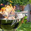 A close-up shot of a barbecue grill with wood burning inside, set in a garden with flowers and greenery. Produktbild-Vorschau